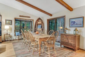 A dining area with a wooden table and chairs, a rustic chest of drawers, and a large window with a view of trees.