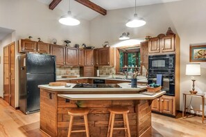Une cuisine avec des meubles en bois, un réfrigérateur noir, un grand îlot central avec des tabourets, des luminaires suspendus et un four à micro-ondes.