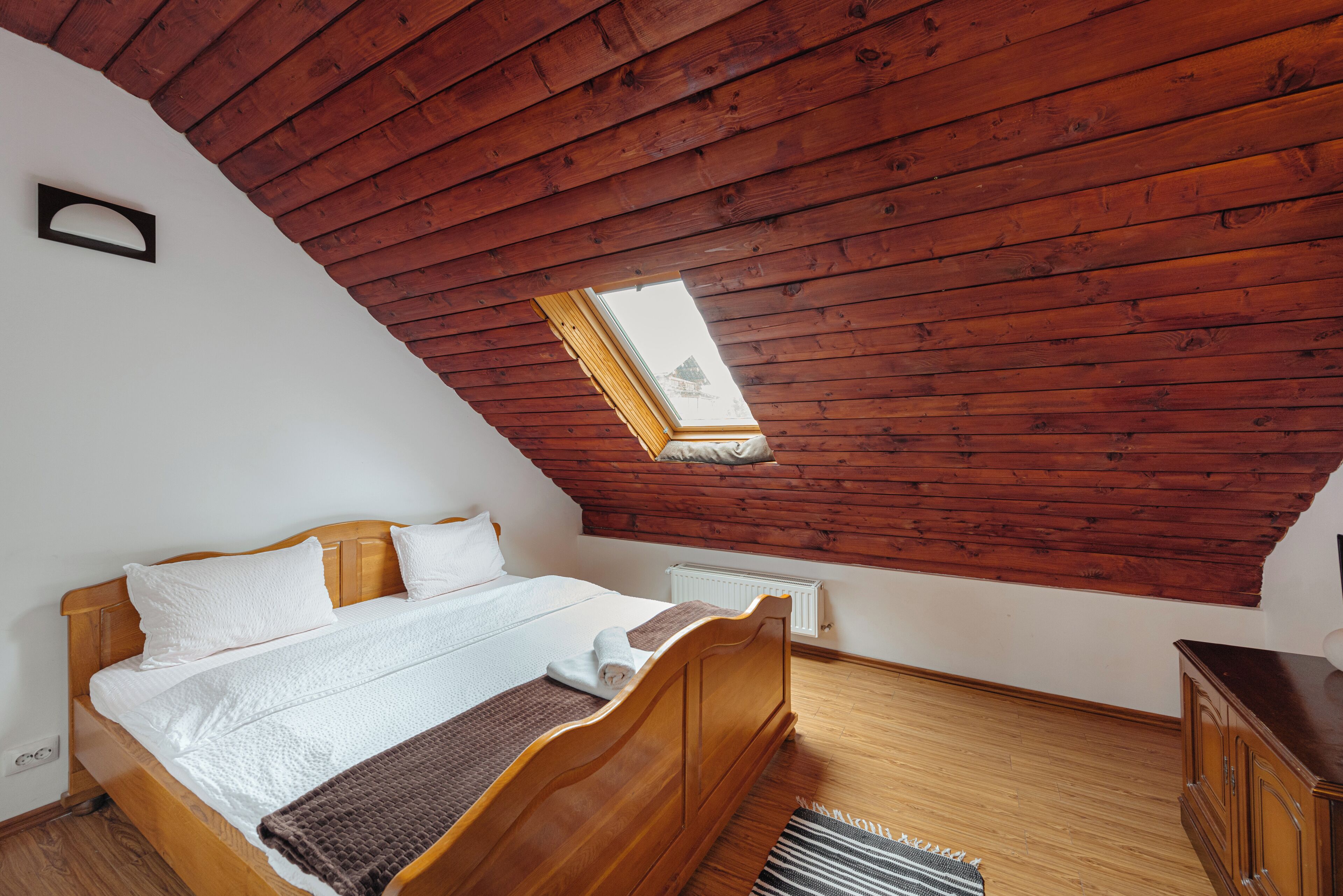 A bedroom with a wooden ceiling, a skylight, a bed with white bedding, and a radiator.