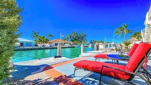 A poolside area with red lounge chairs, a clear blue sky, and a view of the water and buildings in the background.