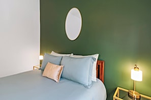 A bed with light blue bedding, a beige pillow, and a wooden headboard. There is a bedside table with a lamp and a mirror on the wall above the bed.