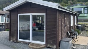 A small, single-story cabin with a sliding glass door, a gravel driveway, and a green building in the background. - Charming Chalet Nestled in Heart of Clarach Bay (Aberystwyth)
