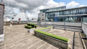Una terraza en la azotea con zonas de estar, un edificio moderno con grandes ventanales y un cielo nublado.