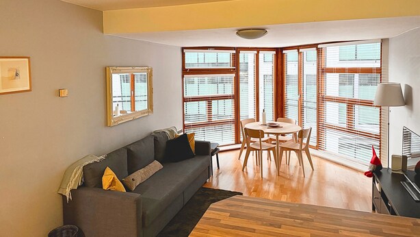 Una sala de estar moderna con un sofá gris, piso de madera y ventanas grandes con persianas.