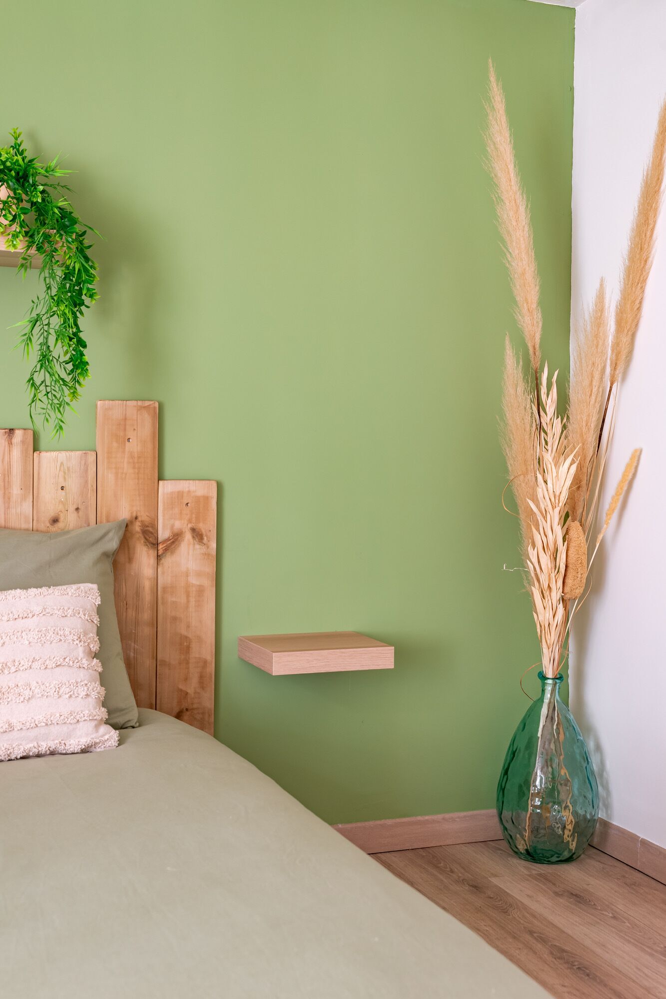 Un dormitorio con cama, pared verde, cabecero de madera, mesita de noche con un jarrón de plumas de pampa y una planta colgante.