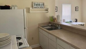 A kitchen with a stove, sink, and white cabinets. There is a window with blinds and a small shelf with kitchen items. - Country farm cottage (Nesmith)