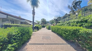 Un chemin pavé, bordé de haies bien entretenues, mène à un bâtiment doté d’un toit en tuiles et de grandes fenêtres.