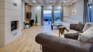 Una sala de estar moderna con chimenea, sofá, televisor y vistas a montañas a través de grandes ventanales.