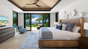 Un dormitorio con una cama grande, un ventilador de techo, vista al océano y una silla junto a la ventana.