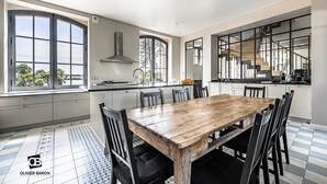 Eine moderne Küche mit einer großen Kochinsel, einem Essbereich mit Holztisch und Stühlen sowie einem Ausblick ins Freie durch große Fenster.