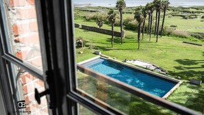 Blick aus einem Fenster auf einen Pool, der auf eine Rasenfläche mit Palmen und ein Gewässer blickt.