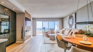 Una sala de estar moderna con sofá, mesa de centro y comedor. Hay un ventanal con vistas al océano.