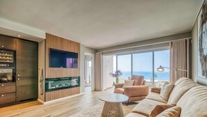Una sala de estar moderna con un sofá grande, una mesa de centro y un televisor montado en una pared de paneles de madera. Se ve el océano a través de amplias ventanas.