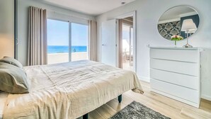 Un dormitorio con cama, ventana grande, espejo y vista a la playa.