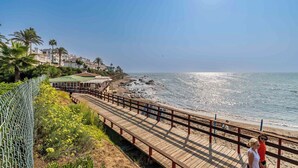 Una pasarela de madera bordeando una playa, con vista despejada al océano y una hilera de edificios al fondo.