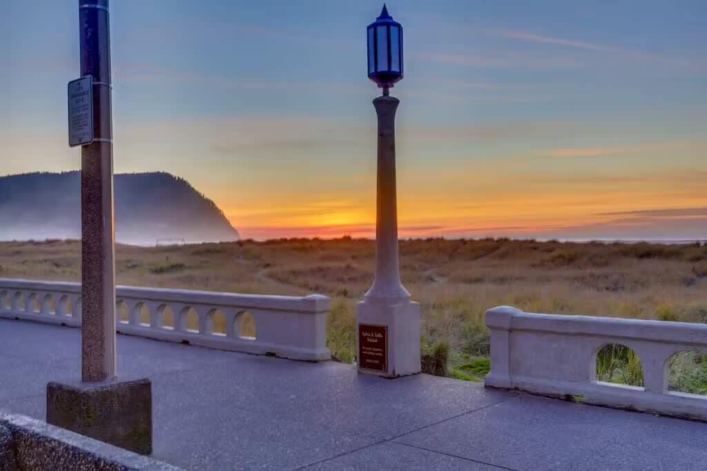 A coastal walkway with a lamppost and a sign, overlooking a sunset over a grassy landscape.