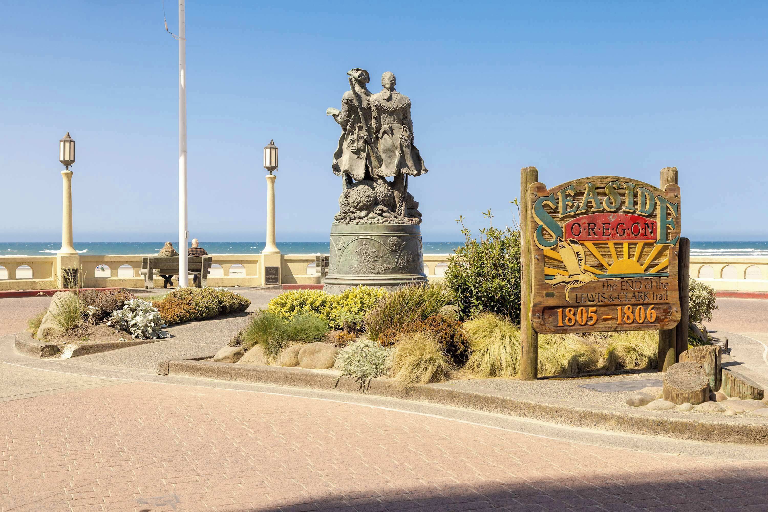 Un monument historique comprenant une statue et un panneau indiquant la fin du sentier de Lewis et Clark, le tout étant entouré de jardins aménagés et offrant une vue sur la côte.
