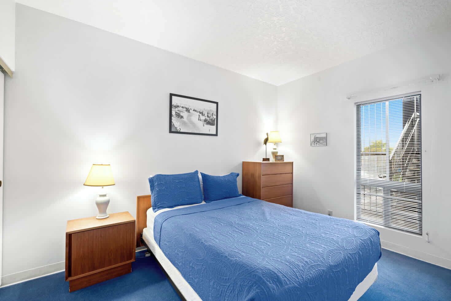 A bedroom with a bed, two bedside tables with lamps, a dresser, a framed picture on the wall, and a window with blinds.