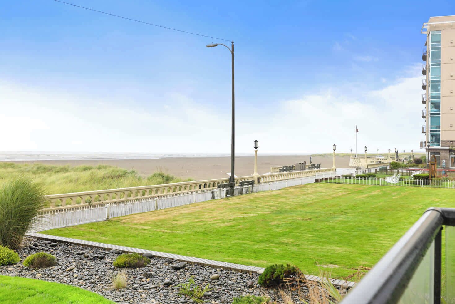 A coastal view with a grassy lawn, a white fence, and a building with balconies.