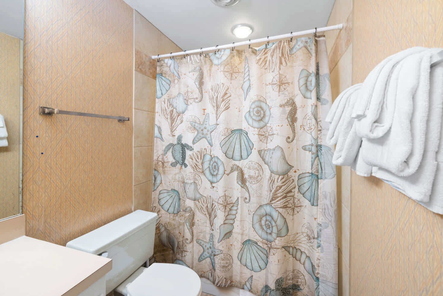 Une salle de bain avec un rideau de douche orné d’un motif représentant des hippocampes et des coquillages, des toilettes et un porte-serviettes avec des serviettes.