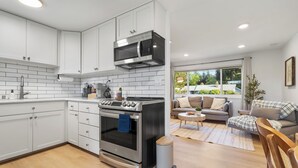 Una cocina moderna con armarios blancos, una estufa de acero inoxidable y un microondas. Hay una zona de comedor con un sofá y una mesa pequeña.