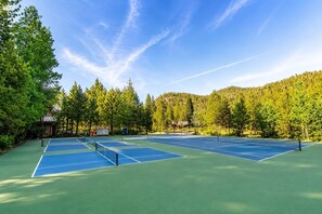 Een tennisbaan met meerdere velden, omgeven door bomen en een bebost heuvel.