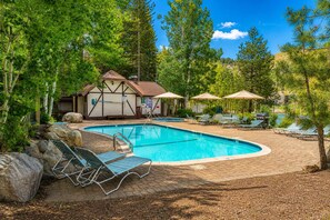 Una piscina con lettini e ombrelloni, circondata da alberi e un edificio con tetto a spiovente.