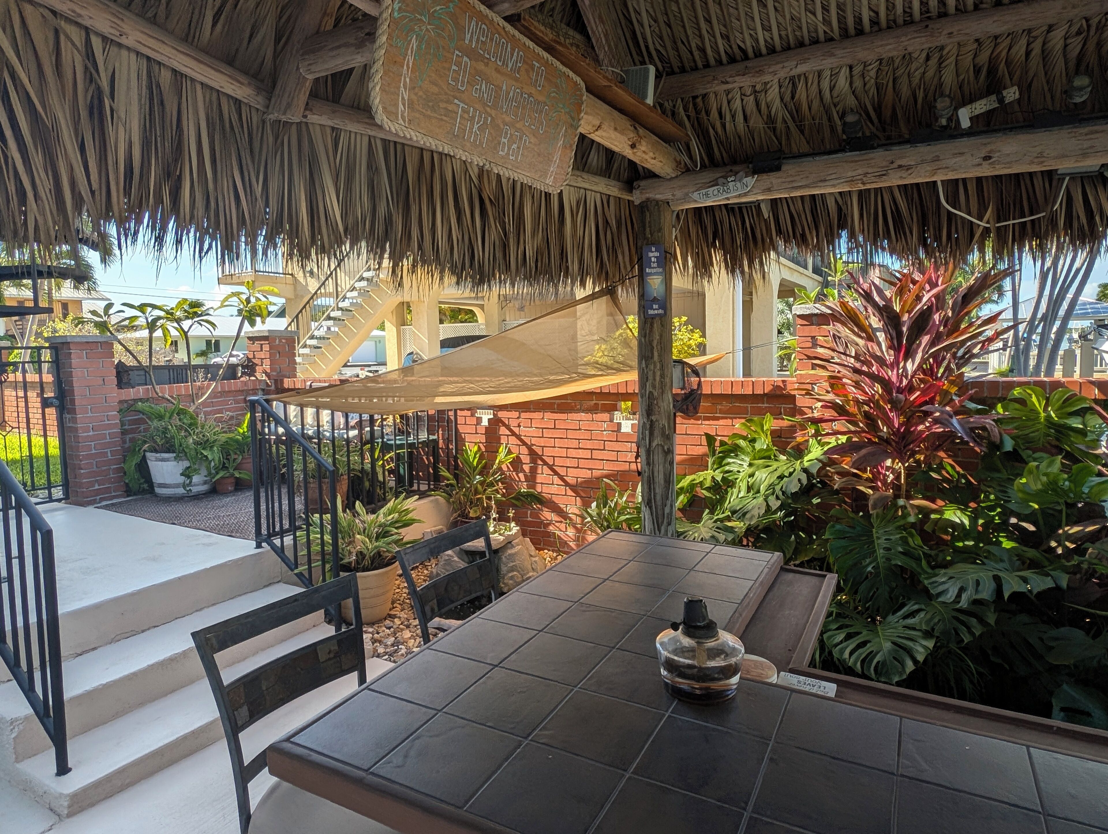 A covered outdoor seating area with a table and chairs, surrounded by potted plants and a brick wall.
