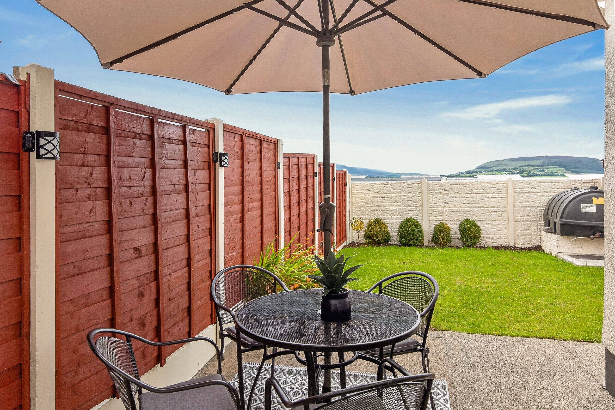 Un patio con tavolo e sedie, un ombrellone grande e vista su una zona verde con un muro in pietra e un barbecue.
