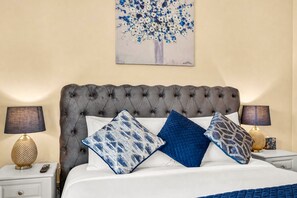 Een slaapkamer met een geborduurde hoofdbord, een witte spreideken en meerdere kussens. Er staan twee nachtkastjes met lampen en een afstandsbediening op een ervan.