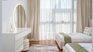Ein modernes Schlafzimmer mit einem weißen Schminktisch, einem runden Spiegel, einem Bett mit grüner Tagesdecke und transparenten Vorhängen.