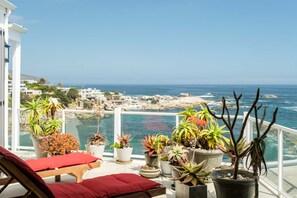 A balcony with potted plants, a red cushion, and a view of the ocean and coastline.