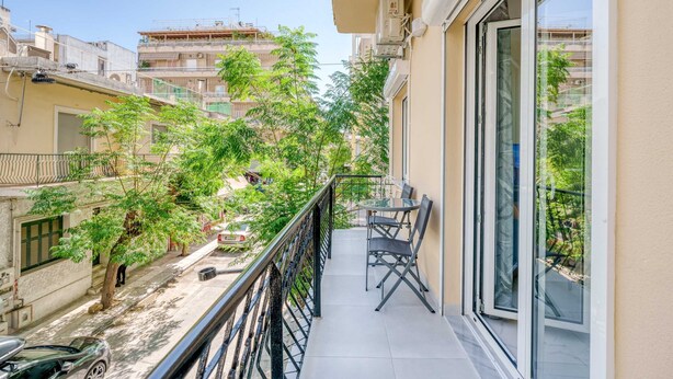 Un balcon avec une table et des chaises, donnant sur une rue bordée d’arbres et sur les bâtiments voisins.