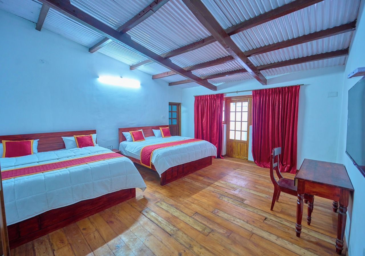 A room with two beds, a wooden floor, a table, and a window with red curtains.