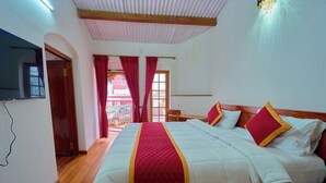 Habitación de hotel con una cama grande, suelo de madera, un televisor montado en la pared y vistas al exterior a través de una ventana con cortinas rojas.