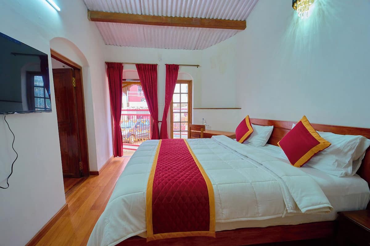 A hotel room with a large bed, wooden flooring, a TV mounted on the wall, and a view of the outside through a window with red curtains.