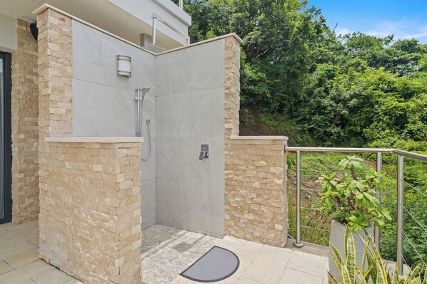 A modern outdoor shower area with stone walls and a metal railing, surrounded by lush greenery.