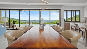 Ein moderner Essbereich mit Holztisch und Stühlen, großen Fenstern mit Blick auf das Meer und die Hügel, sowie eine Küchenzeile mit Bar.