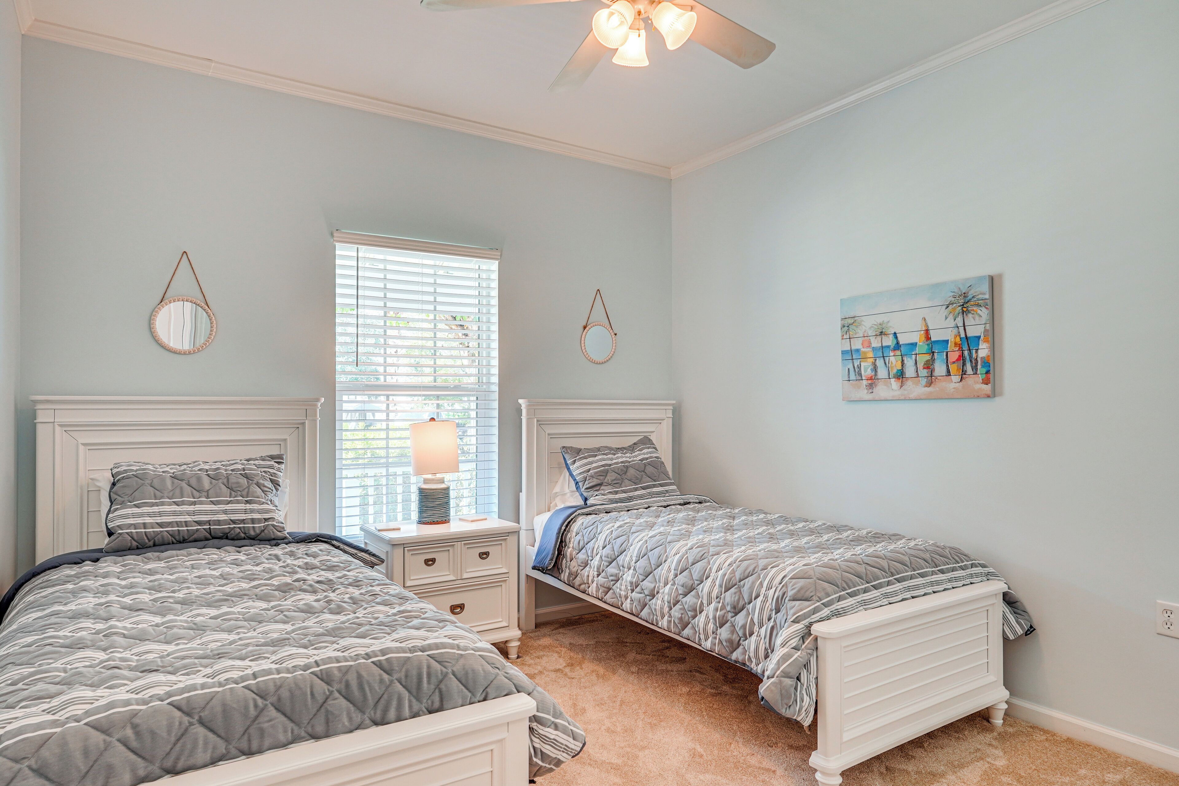 A bedroom with two beds, a nightstand, a ceiling fan, a window with blinds, and a beach-themed painting on the wall.