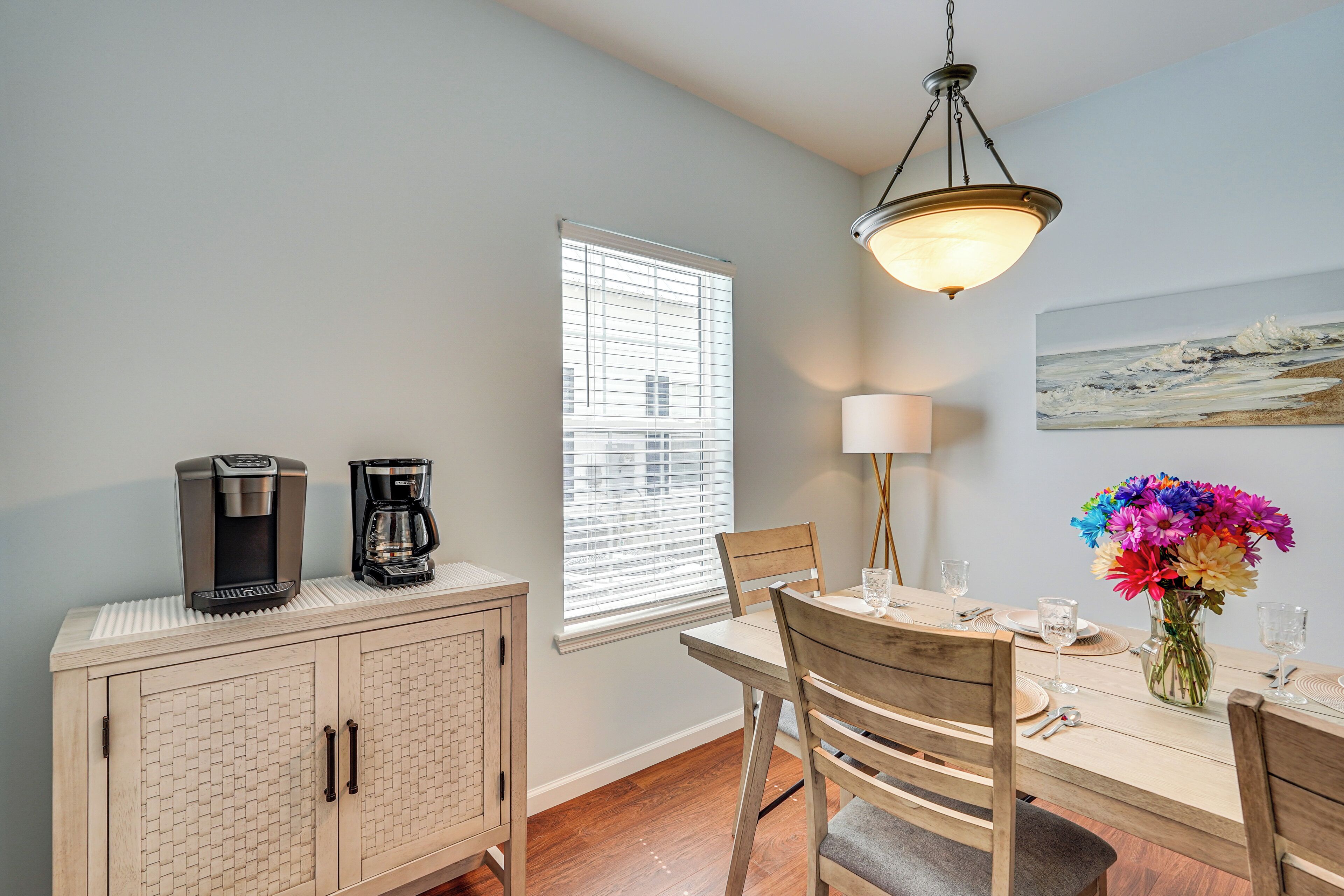 A dining area with a wooden table, chairs, a coffee maker, a lamp, and a vase of flowers.