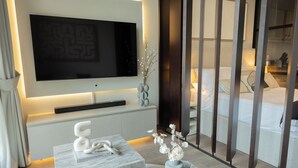 A modern hotel room with a large flat-screen TV mounted on the wall, a marble coffee table, a vase with flowers, and a bed with a wooden headboard. - WelHome - LOCI Residences (Dubai)