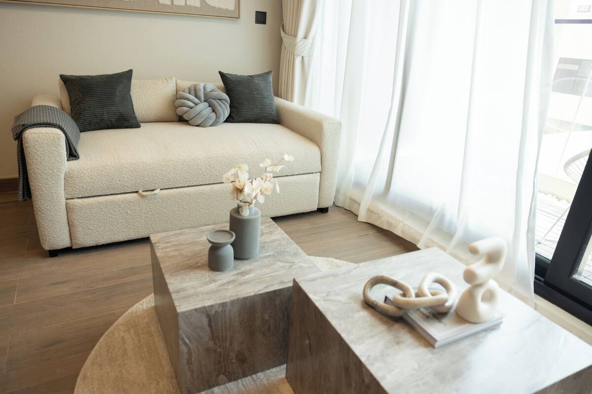 A modern living room with a beige sofa, a wooden coffee table, and a view of the outdoors through large windows.