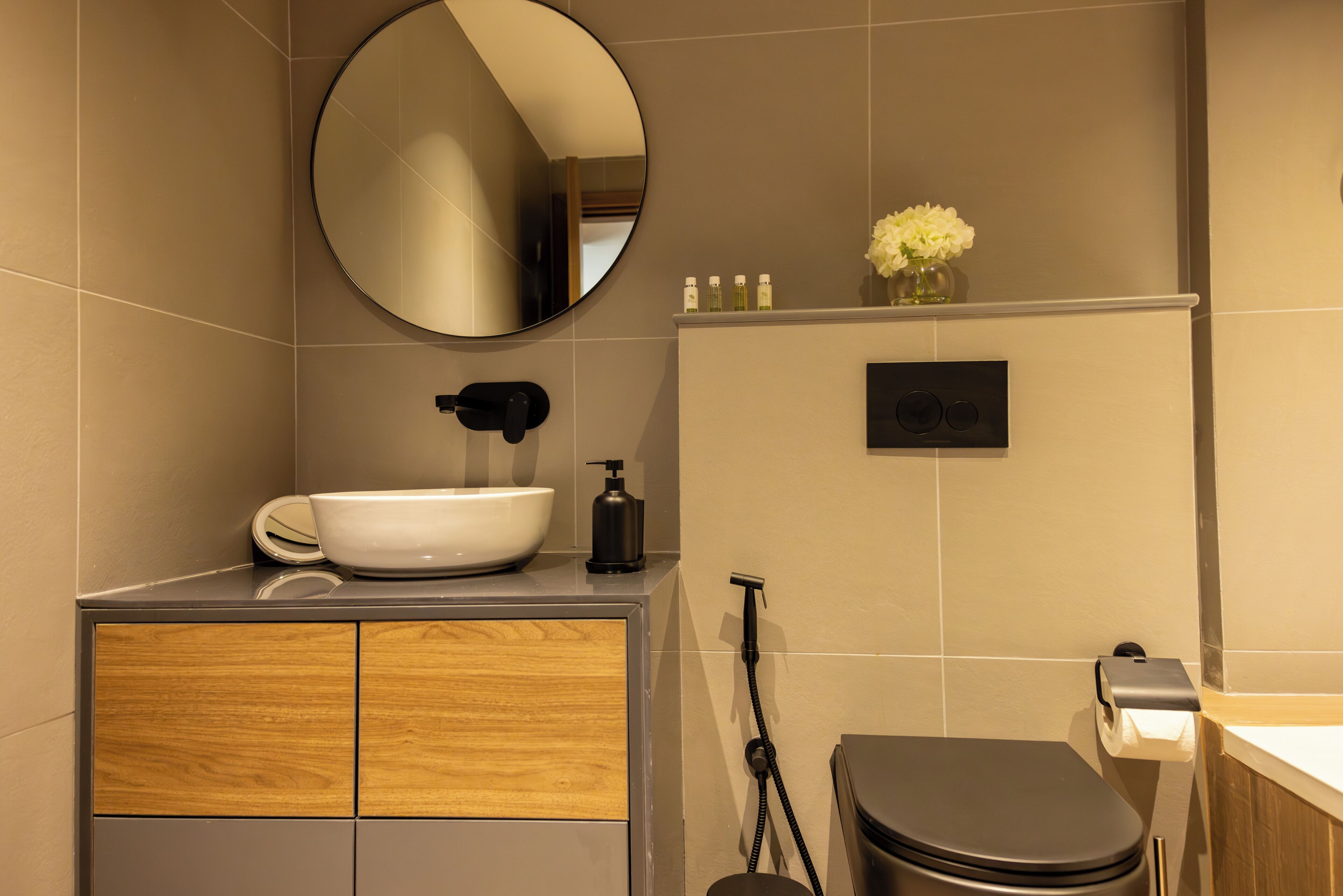 A modern bathroom with a round mirror, a white sink, a wooden cabinet, a toilet, and a small vase with flowers on the wall.