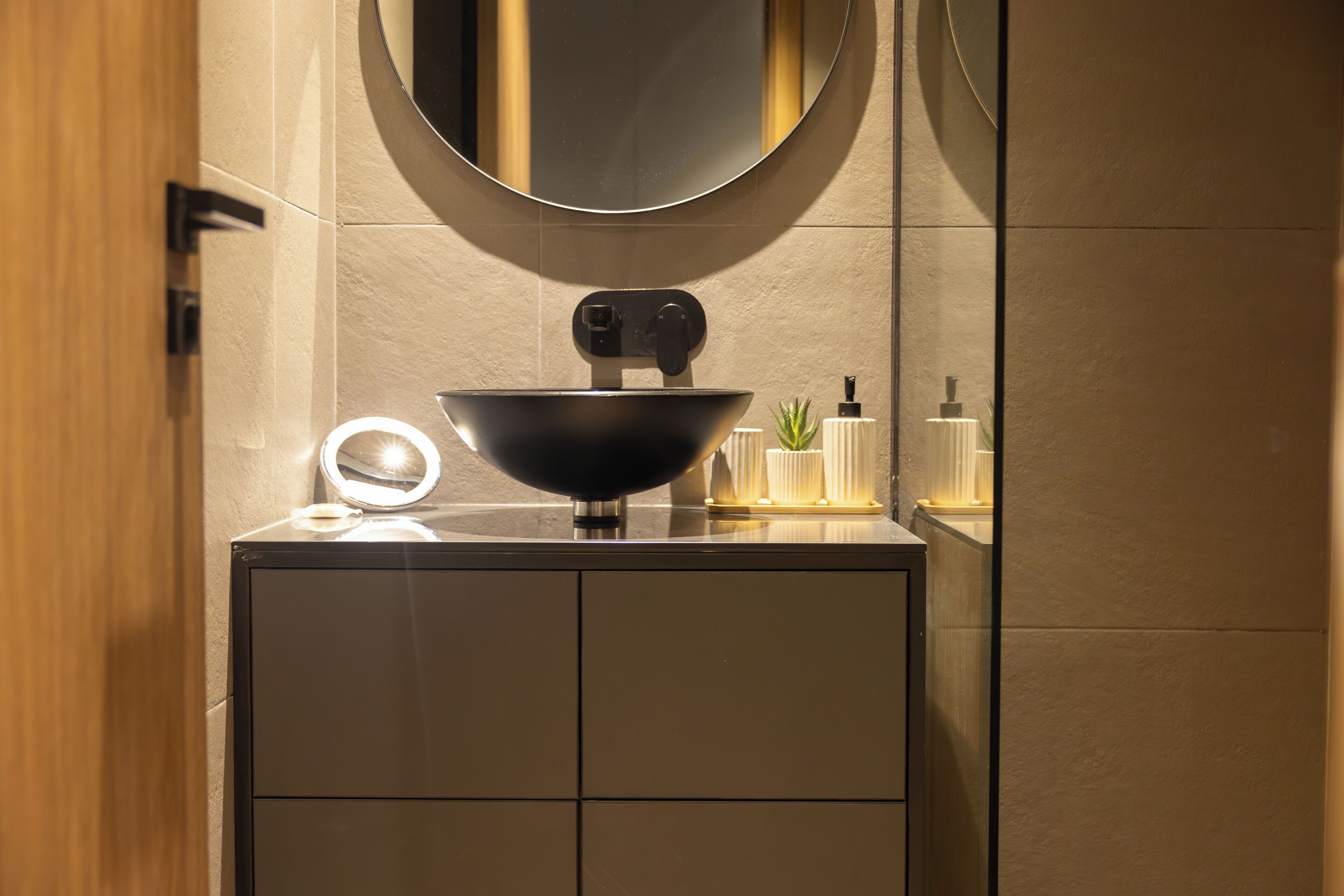 Un baño moderno con un lavamanos negro, un espejo redondo y una planta pequeña en la encimera.