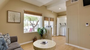 A living room with a round coffee table, a gray sofa, a large window with blinds, a potted plant, and a laundry area with a washing machine. - A-frame w/ Patio & Mtn Views: Arizona Retreat (Apache Junction)