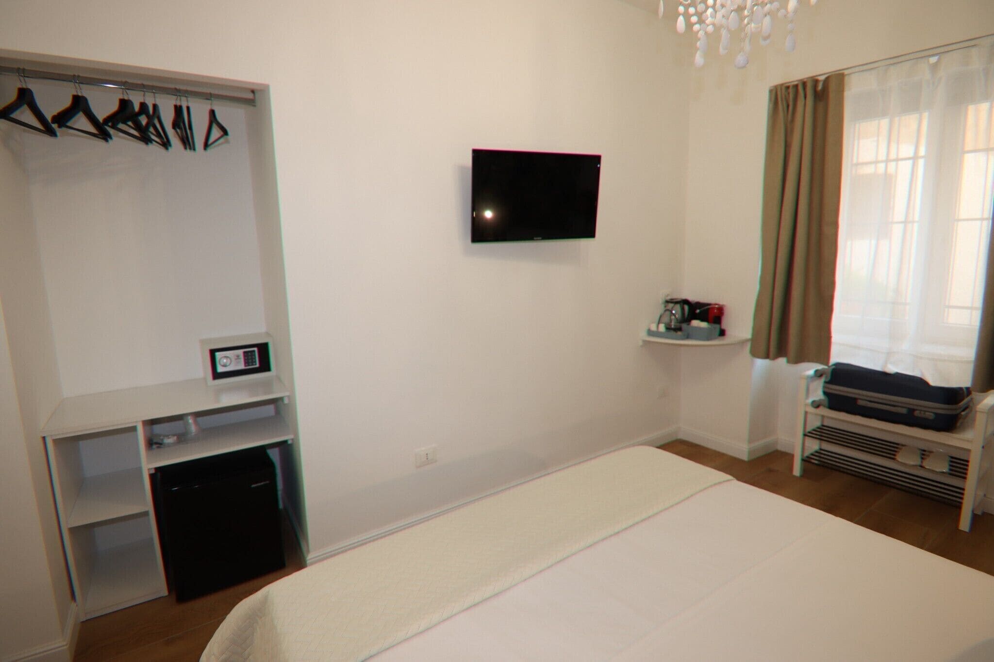 A hotel room with a bed, a TV mounted on the wall, a coat rack with hangers, a small shelf with toiletries, and a window with curtains.
