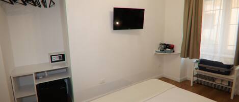 Habitación de hotel con cama, televisor montado en la pared, perchero con ganchos, estante pequeño con artículos de aseo y ventana con cortinas.