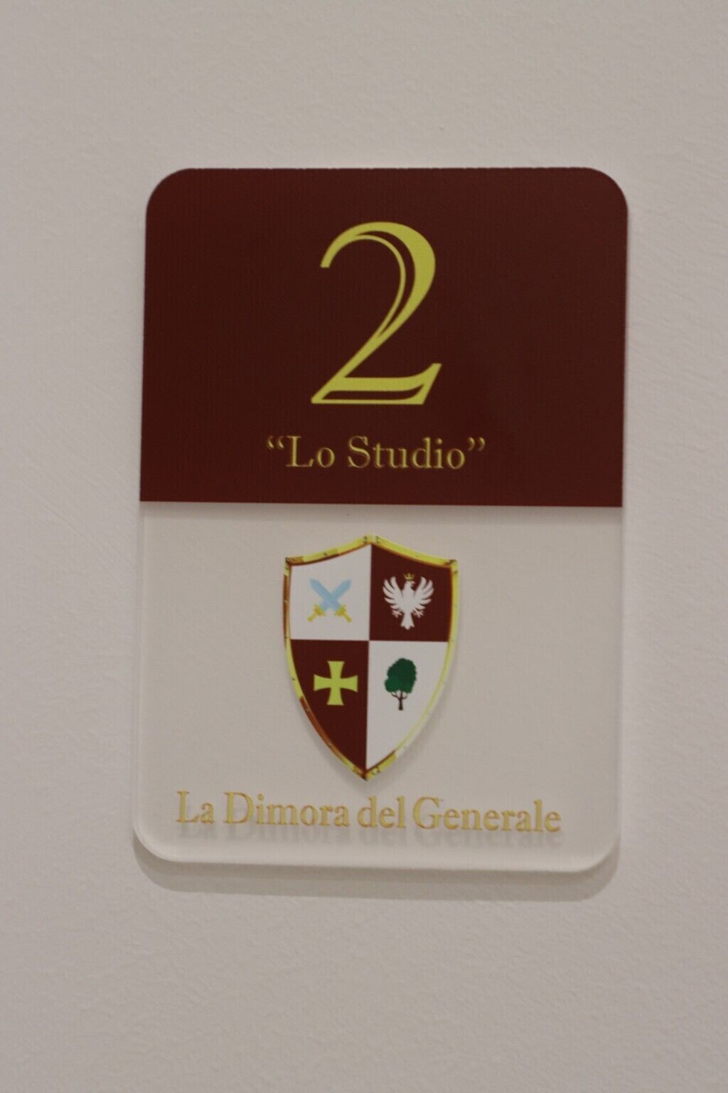 A badge with the number 2 and the text "Lo Studio" on a maroon background, and a shield with various symbols on a white background below it.