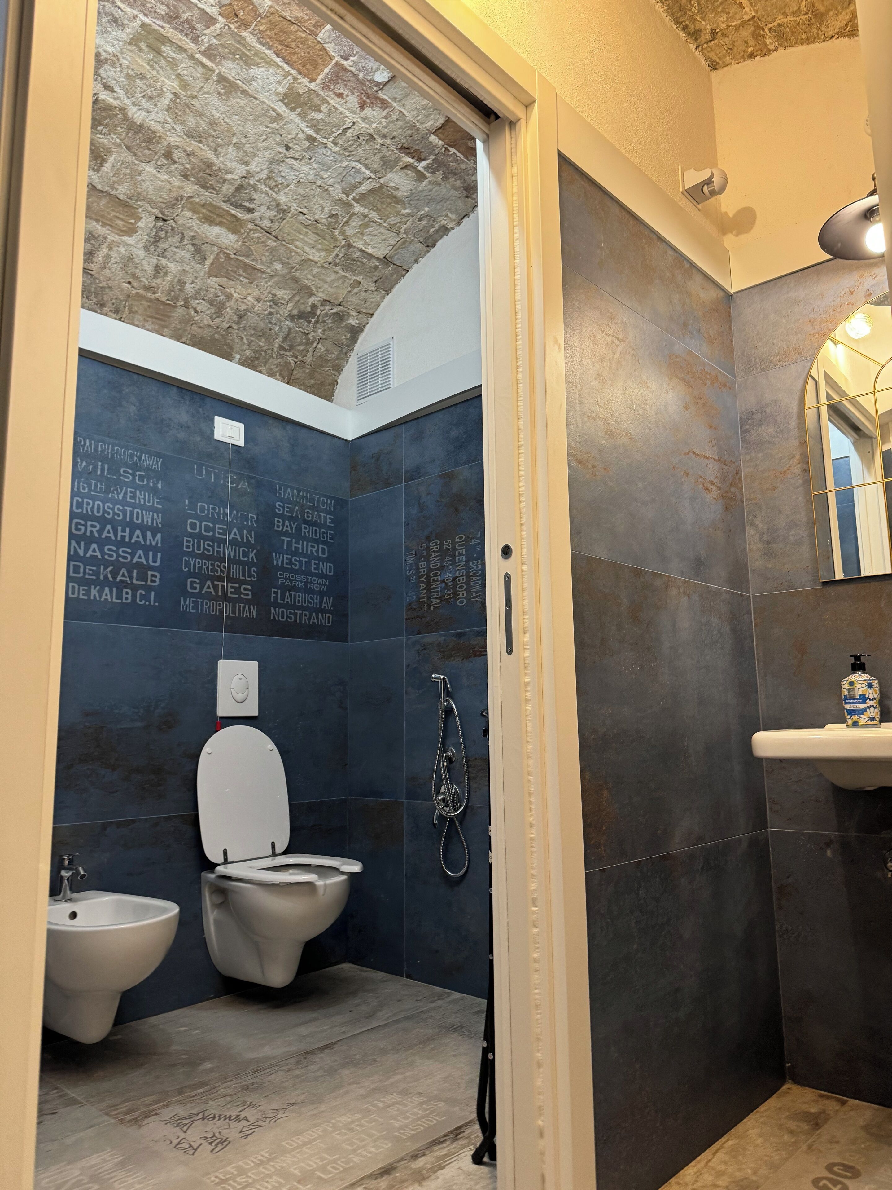 A bathroom with a stone wall, a toilet, bidets, and a sink with a mirror.