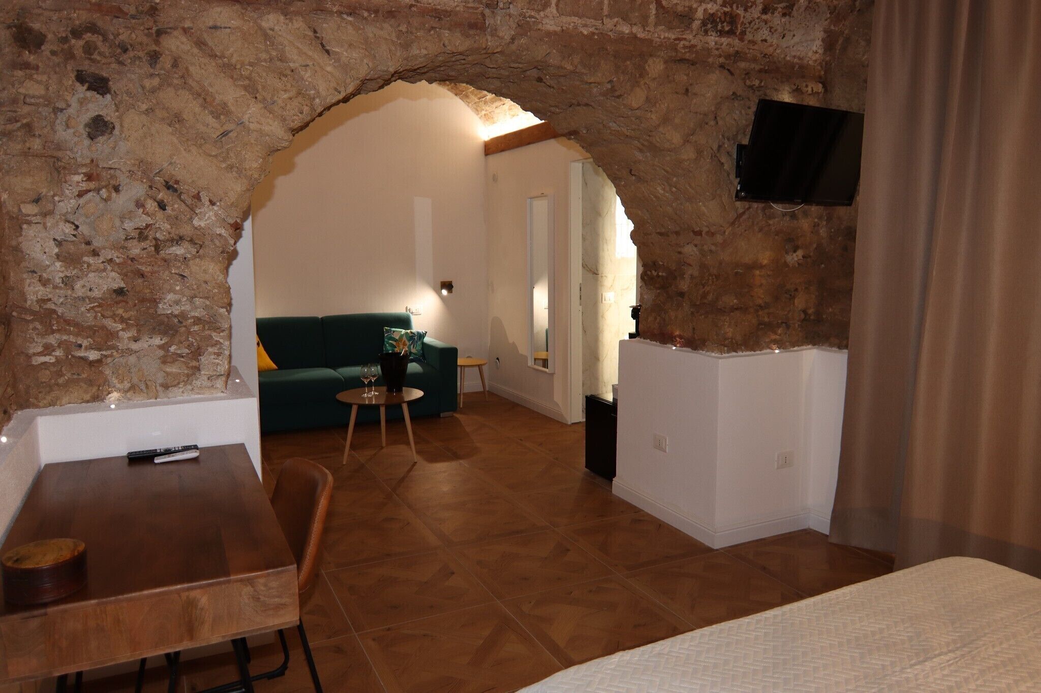Ein Zimmer mit einem Steingewölbe, einem grünen Sofa, einem kleinen Tisch mit einem Weinglas und einem an der Wand befestigten Fernseher.